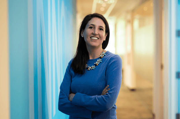 Cristina smiling wearing a white blouse and black blazer with a light blue background banner